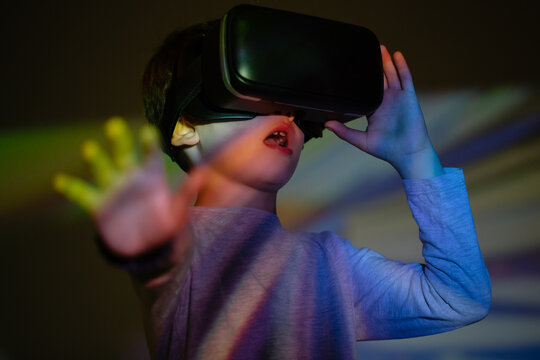 Cute boy wearing VR glasses and exploring virtual reality with excited face expression while standing under colorful projection