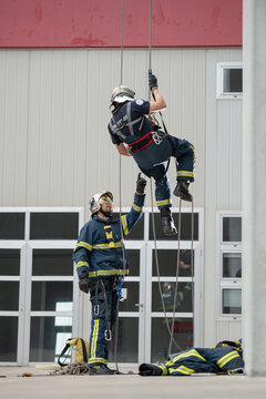 Brave fireman climbing rope under control of skilled male colleague near building in training center