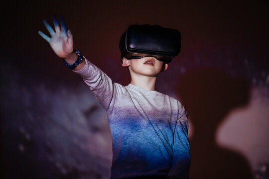 Cute boy wearing VR glasses and exploring virtual reality while standing under colorful projection