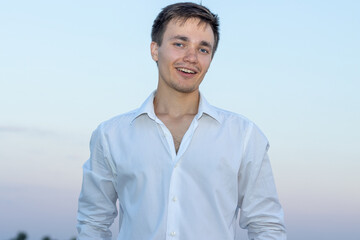 Portrait of a happy young man in a white shirt on the sunset sky background.