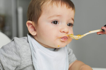 Adorable little baby sitting on high chair and eating fresh avocado from spoon held by crop anonymous mother