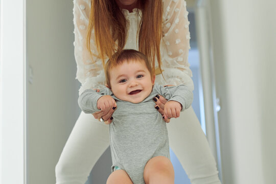 Cute cheerful toddler in gray bodysuit held by crop unrecognizable mother smiling and looking at camera  while having fun together at home