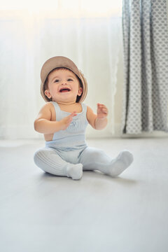 Adorable cheerful little baby wearing hat and looking away while sitting on floor in light nursery at home