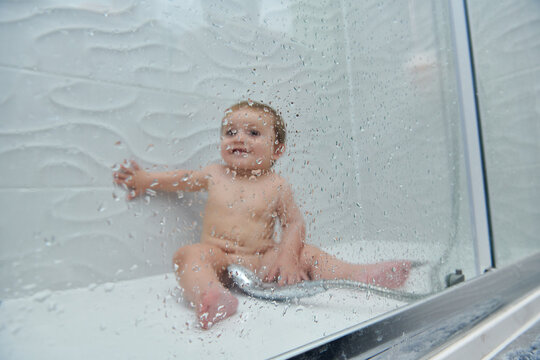 Cute cheerful little toddler sitting on floor of shower cabin and enjoying water procedure during bathing time at home