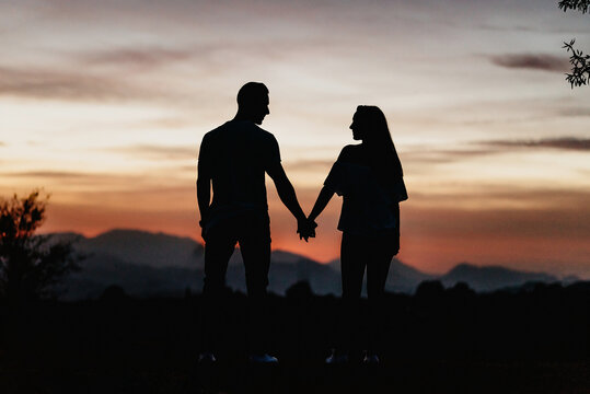 Back view silhouettes of unrecognizable man and woman holding hands and looking at each other while standing on hill during beautiful sunset in mountains