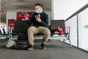Focused male traveler in casual wear and face mask using smartphone to take selfie holding passport and boarding pass while sitting on chairs with backpack in airport waiting area during coronavirus epidemic