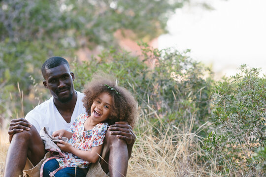 Positive black man in casual clothes sitting with cute little daughter on laps and leaning on tree trunk while spending sunny summer day in forest