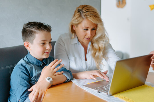 Content mother in white blouse and cute positive grade schooler son in denim sitting at table together and browsing modern netbook in light living room