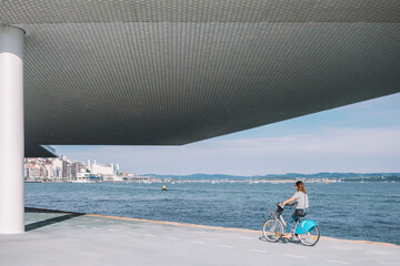 Side view of female in casual wear riding bike near modern building and waterfront during sunny day