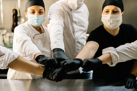 Content cooks team wearing uniform face masks and gloves striking bargain with fists while working in modern restaurant kitchen during coronavirus epidemic