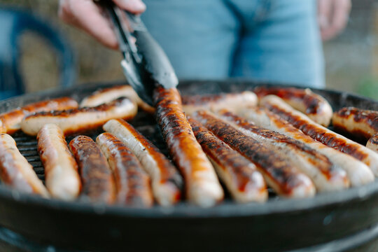 Crop anonymous person in jeans frying delicious beef sausages on grill and using tongs while having barbecue picnic on backyard