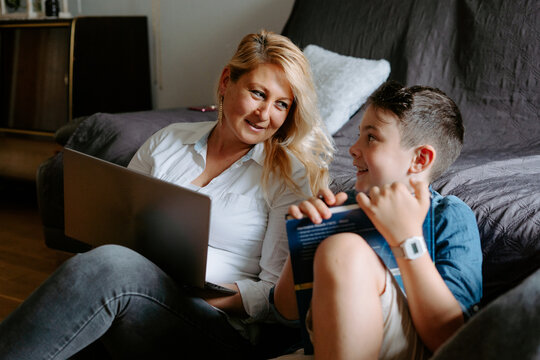 Happy smiling mother with glass of cocktail using netbook while sitting near content son reading book while spending free time together