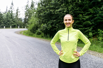 A portrait of attractive positive woman in sportswear outdoors, she stands with hands on hips position and looks at camera