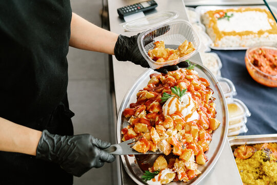 From above crop anonymous waitress wearing black latex gloves putting delicious baked sweet potatoes into plastic container for takeaway
