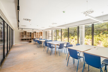 Modern style office interior with square tables and blue stools on floor and wooden shelves on wall with large window and lamps on ceiling