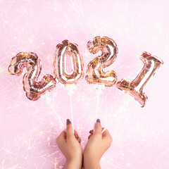 Happy New Year 2021 celebration. Female hands holding golden balloons in form of numbers with bright sparklers on pink background. Flat lay, top view