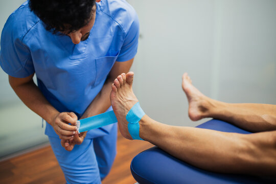 Top view of crop unrecognizable chiropractor in blue uniform attaching kinesio tape on foot of faceless man lying on examination couch in clinic
