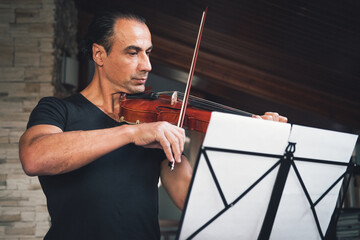 Talented Hispanic male violinist playing violin and looking at music sheets during rehearsal