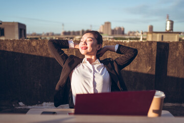 Young Asian female employee in formal wear sitting at table with laptop and cup of coffee on rooftop terrace and relaxing after remote work