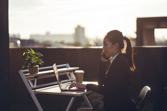 Side view of young Asian business lady in formal outfit talking on smartphone and using laptop while working remotely on rooftop terrace in city