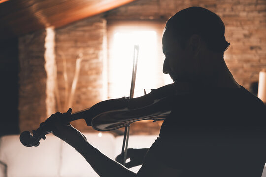 Side view of silhouette of male musician playing violin and looking at music sheets while practicing in living room