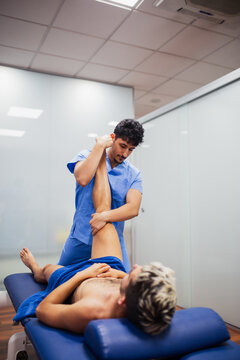 Osteopath in blue uniform examining knee of unrecognizable slim male patient with dyed hair lying on examination table in clinic