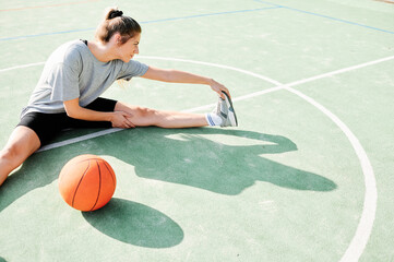 High angle view of young female basketball player sitting on court near ball and doing stretching exercise before game