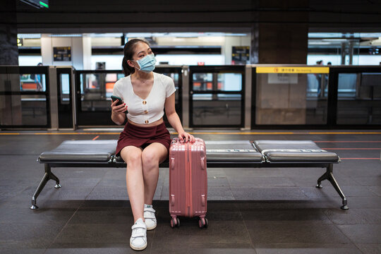 Young concentrated Asian lady wearing mini skirt and face mask browsing mobile phone while sitting on bench with suitcase in train station