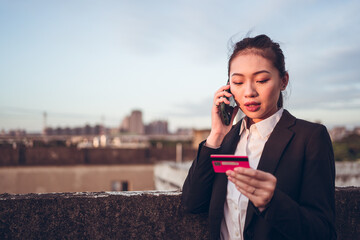 Serious young Asian businesswoman in formal wear holding credit card and having phone conversation with bank customer service while standing on rooftop of city building