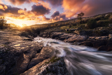 Picturesque scenery of rapid creek flowing through rough stony terrain under colorful cloudy sunset sky