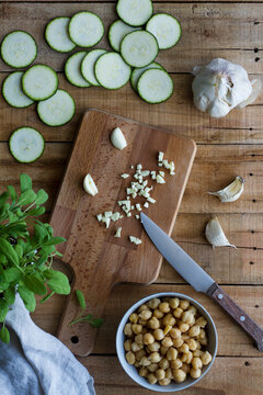Top view of bowl with cooked chickpeas placed on wooden table near chopped garlic and sliced zucchini with fresh herbs during preparation of healthy vegan food