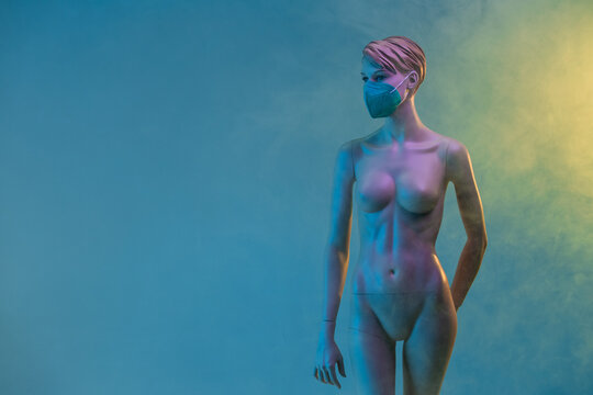 Plastic female mannequin in medical mask illuminated by colorful lights in studio with smoke on blue and yellow background