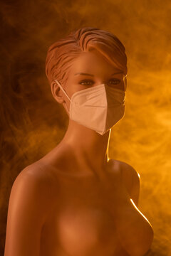 Plastic female mannequin in medical mask illuminated by colorful lights in studio with smoke on orange background
