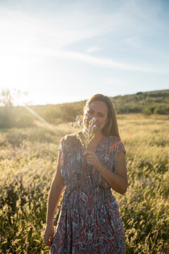 Smiling female wearing trendy dress and straw hat standing on meadow during sunny day and enjoying summer holding fresh perfumed flower