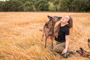 Female with closed eyes cuddling gently purebred dog with smooth coat while sitting in field with faded grass near forest in countryside under sky with clouds