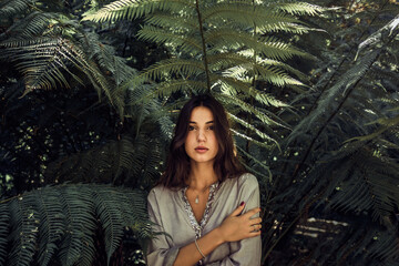Tranquil female in summer outfit standing in shade of green leaves of tree on sunny day and looking at camera