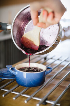 Crop anonymous housewife with wooden spoon pouring tasty freshly made blueberry jam from pan into ceramic pot while preparing breakfast at home