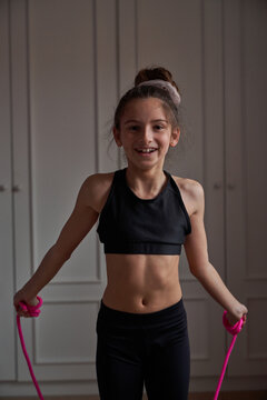Teen slender gymnast in black sportswear jumping rope while training in own room at home looking at camera