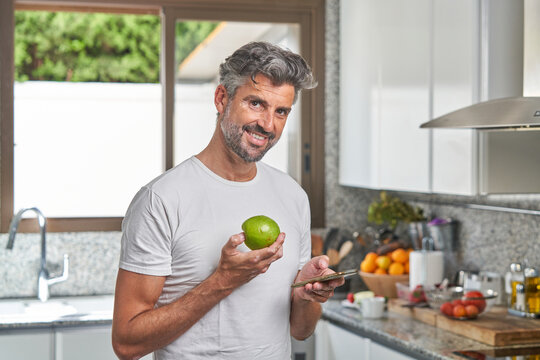 Cheerful Mature Male In Casual T Shirt Standing In Kitchen With Green Apple And Reading Message On Smartphone During Breakfast In Morning