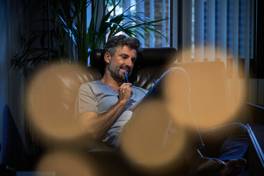 Delighted mature male in casual clothes resting in comfortable armchair and taking notes in notepad