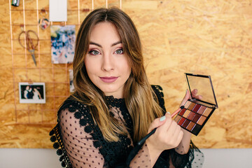 Charming female influencer in elegant wear standing with palette of eyeshadow and cosmetic brush while looking at camera making a tutorial