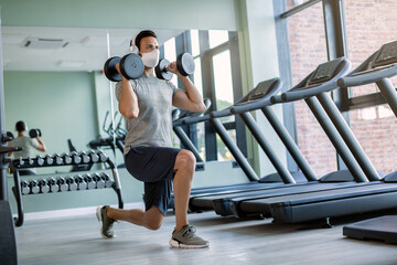 Fototapeta na wymiar Male athlete wearing face mask while exercising with hand weights in lunge position in a gym.