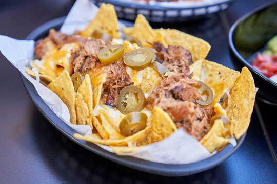 From above of black basket with corn nachos topped with pulled meat in sauce and jalapeno slices in cafe