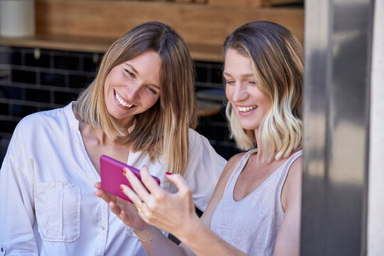 Expressive girlfriends grimacing while taking selfie with smartphone in cafe sitting near opened window