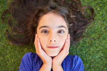 From above of astonished preteen girl with wavy brown hair keeping hands on cheeks and looking at camera while lying on green grass