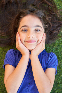 From above of happy preteen girl with wavy brown hair keeping hands on cheeks and looking at camera while lying on green grass