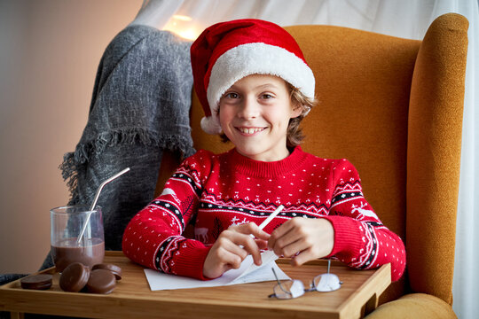 Cheerful child in Santa hat sitting in armchair with pencil in front of table tray with glass of chocolate drink and drinking tube near choco pies and looking at camera