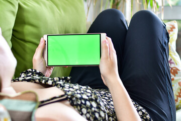 Young woman lying on the sofa, watching content on her mobile with green screen.