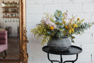 Beautiful bouquet with various flowers including goldenrod and craspedia flowers with green eucalyptus branches arranged in ornamental ceramic pot placed on small table against white brick wall with mirror in creative floristry studio
