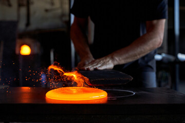 Faceless man working with liquid hot glass while blowing art elements in dark workshop